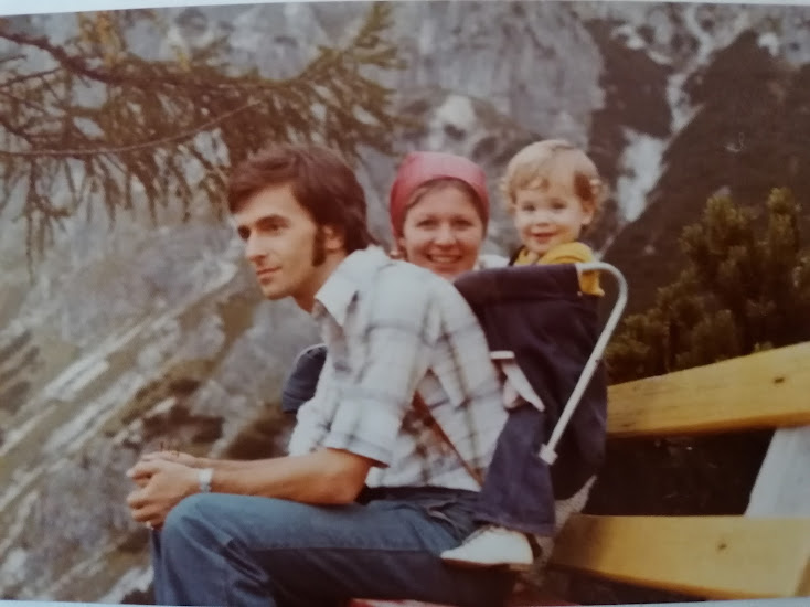 Meine Eltern mit mir als Kleinkind auf einer Bank sitzend auf einem Ausflug in den Bergen. Kin in einem Tragegestell am Rücken.